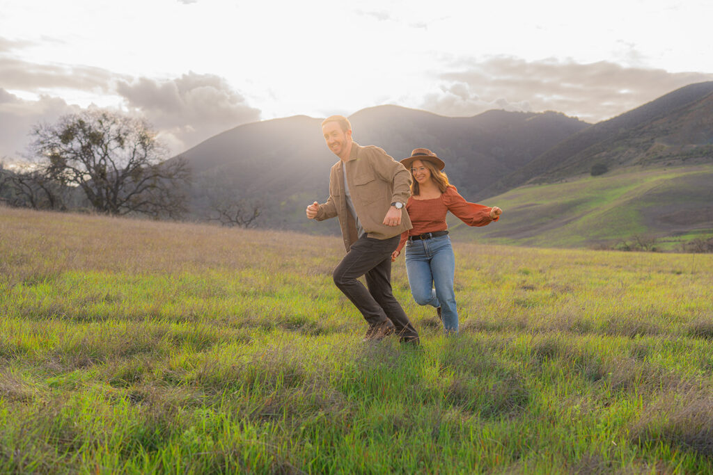 Couple playfully chasing each other through an open field near a rustic water tower and Mt. Diablo in the Bay Area.