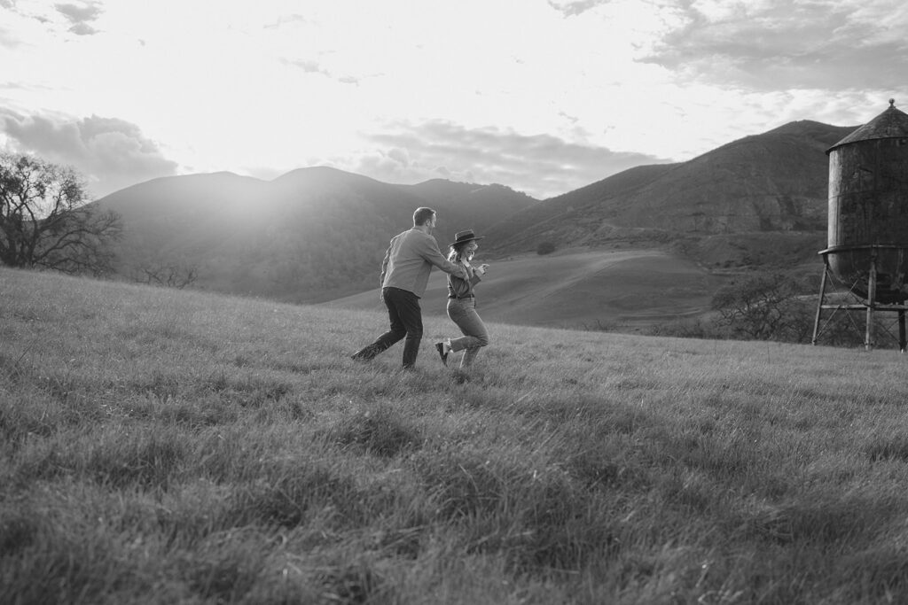 Couple playfully chasing each other through an open field near a rustic water tower and Mt. Diablo in the Bay Area