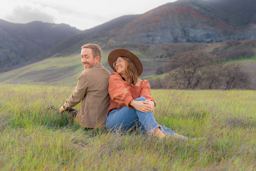 Couple sitting together, one wearing a hat, in an open field near a rustic water tower and Mt. Diablo in the Bay Area.